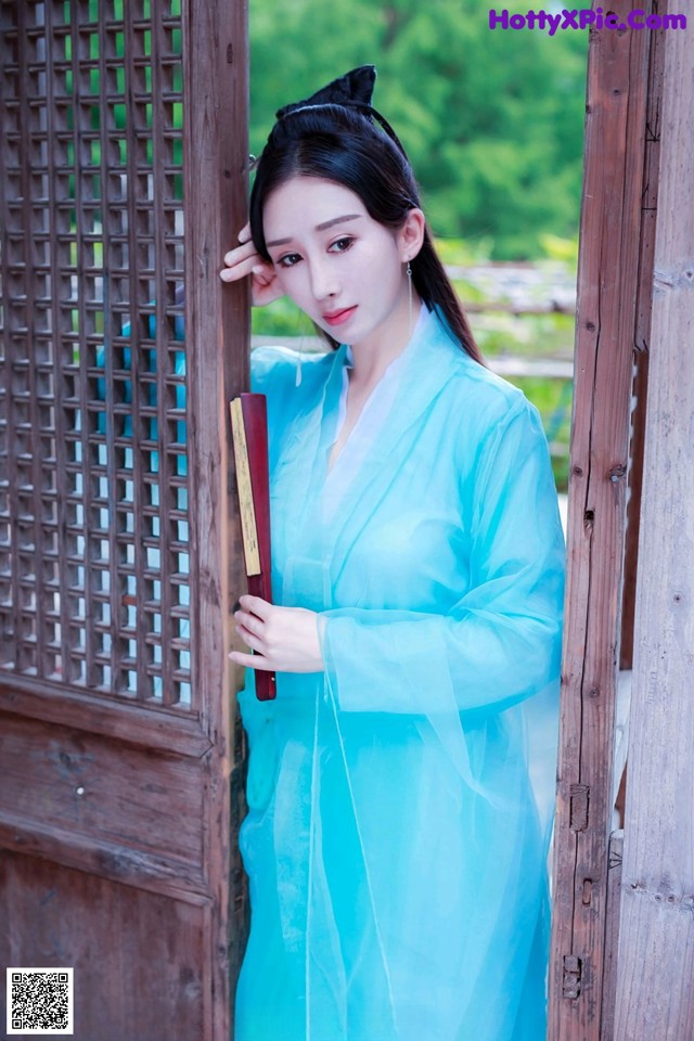 A woman in a blue dress leaning against a wooden door.