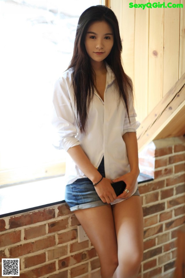 A woman in a white shirt and denim shorts leaning against a brick wall.