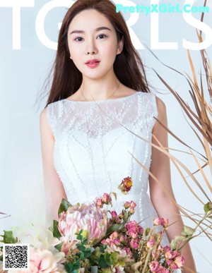 A woman in a white dress sitting on a wooden bench surrounded by flowers.