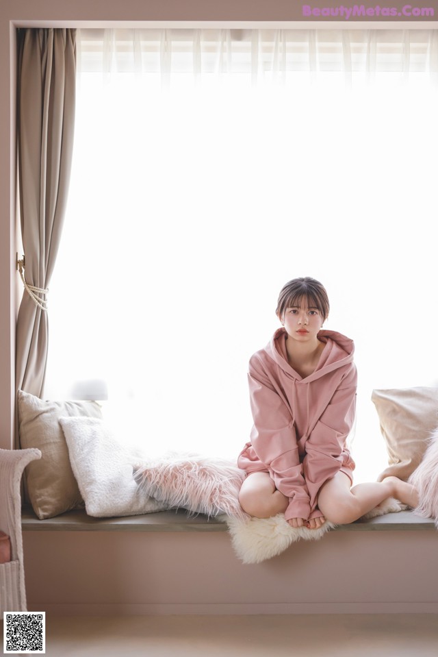 A woman sitting on a window sill in a pink robe.