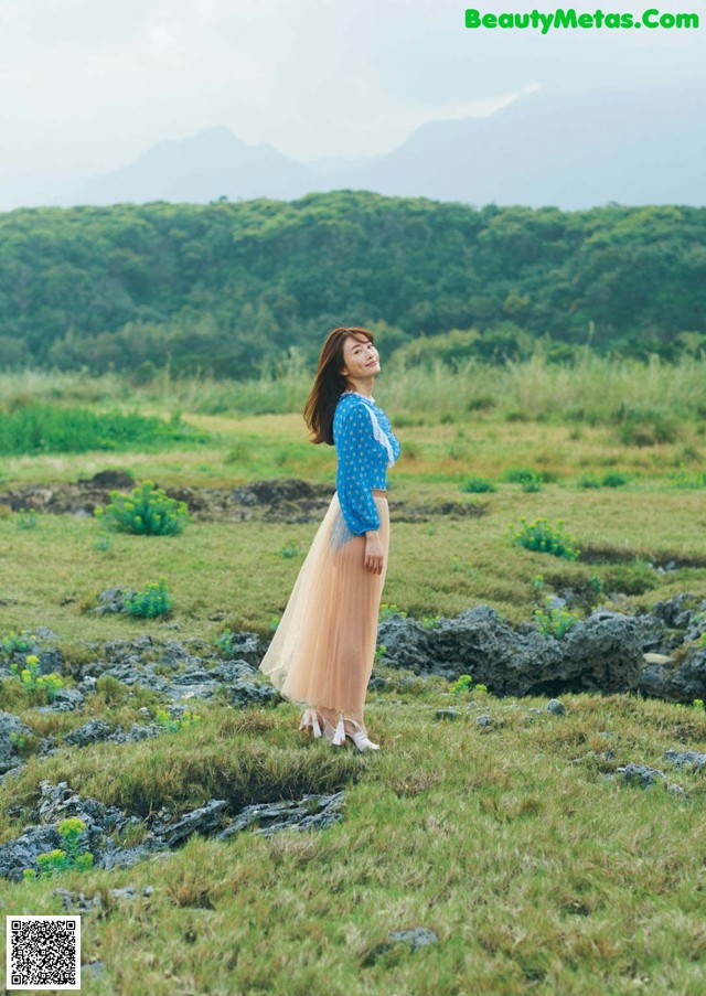 A woman standing in a grassy field with mountains in the background.