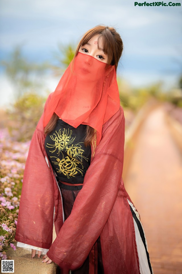 A woman wearing a red veil standing on a dirt road.